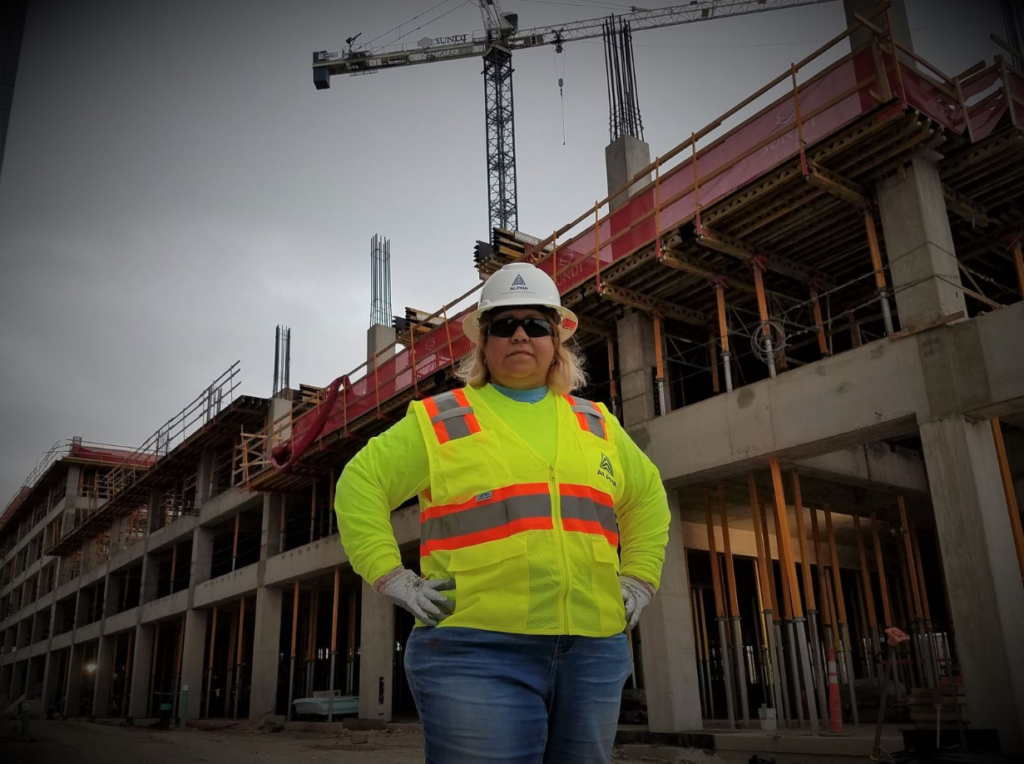 Alpha Insulation & Waterproofing technician in yellow safety vest and hard hat, with hands on hips, standing in front of a building under construction.